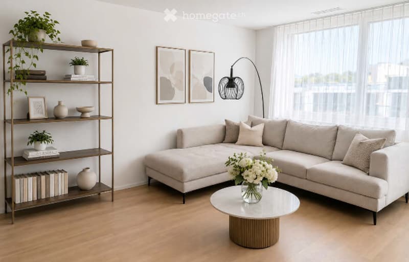 Modern living room with a light grey sectional sofa, a round wooden coffee table with a flower arrangement, a tall wooden shelf with books and decorative items, and large windows with sheer curtains.