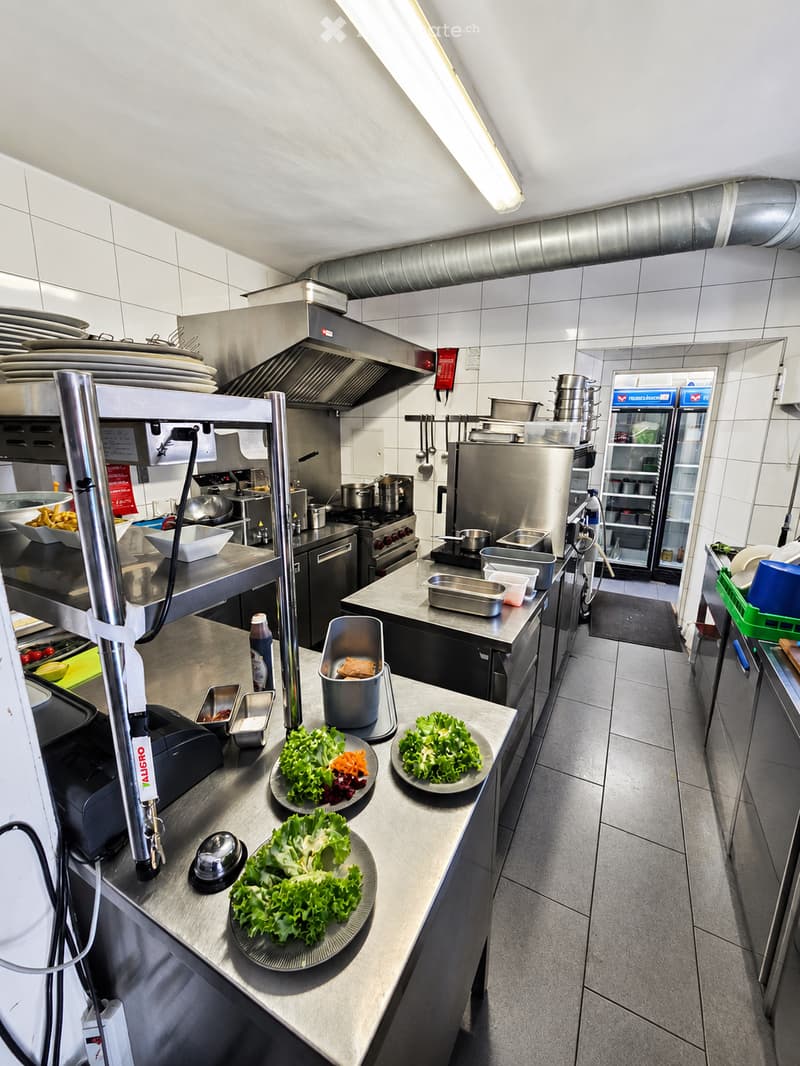 Commercial kitchen with stainless steel appliances, including a refrigerator, oven, stove, and dishwasher. Countertops display plates of lettuce and other food items, along with a cash register and various kitchen tools.