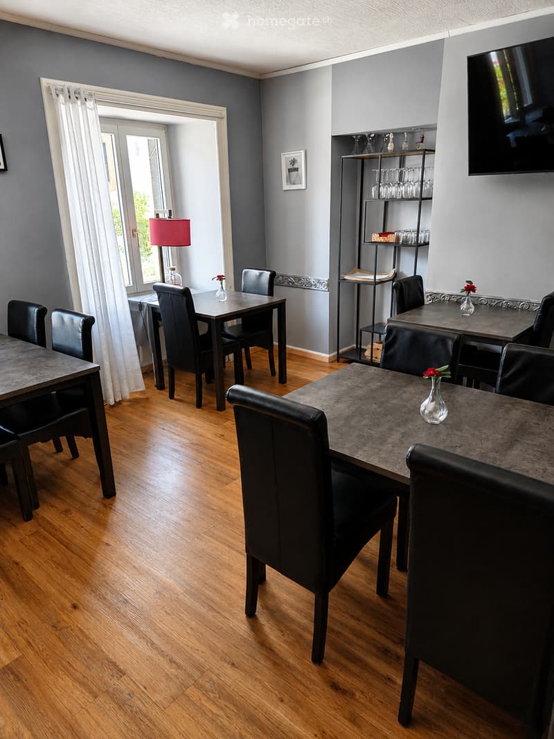 Modern dining area with wooden flooring, multiple tables and chairs, a lamp, a shelf with glassware, and a television mounted on the wall.