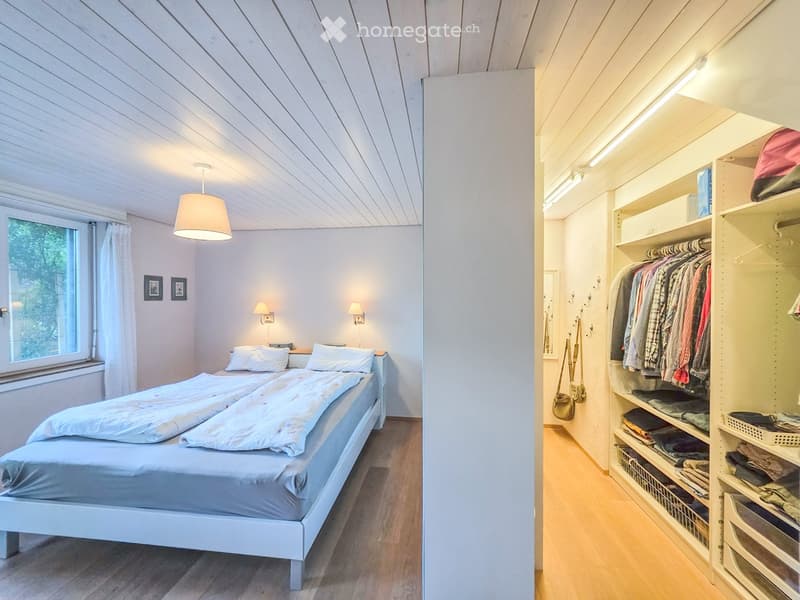 Cozy bedroom with a double bed, nightstand, wall-mounted lamp, and a built-in wardrobe with hanging clothes and storage baskets.