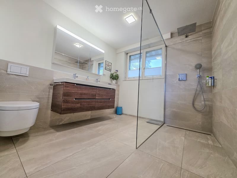 Modern bathroom with beige tiles, a wooden vanity with a sink, a toilet, a large mirror, and a glass-enclosed shower with a handheld showerhead.
