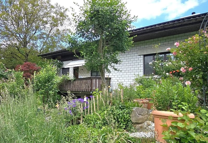 A well-maintained garden with a variety of plants, flowers, and trees surrounding a modern house with a tiled exterior and a wooden balcony.