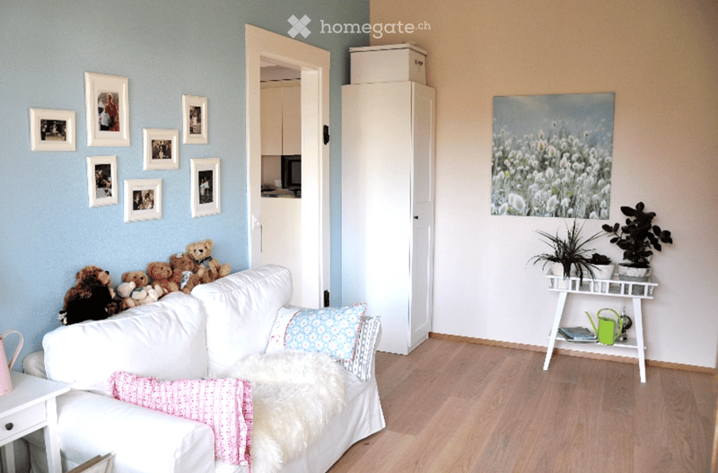 A cozy living room with a white sofa adorned with colorful pillows and a furry throw, surrounded by stuffed toy bears. A small table holds potted plants and a green watering can, with picture frames decorating the walls and a view into an adjacent kitchen
