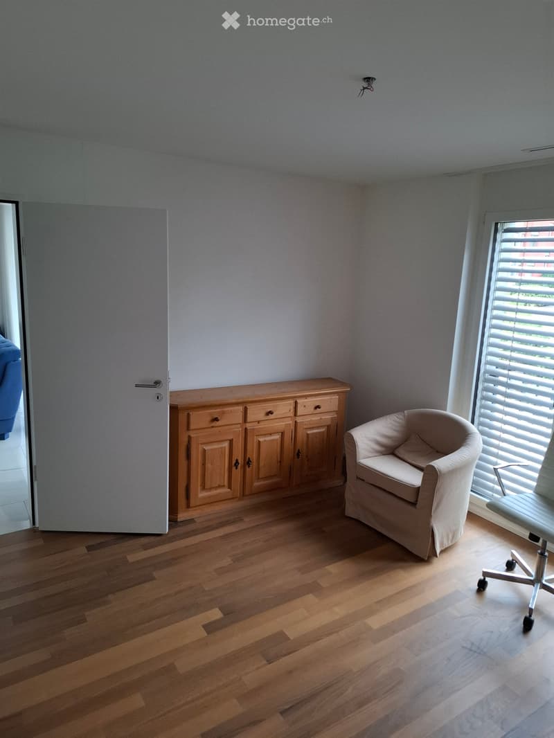 A furnished room with wooden flooring, a light-colored armchair, a wooden cabinet, and an office chair near a window with blinds.