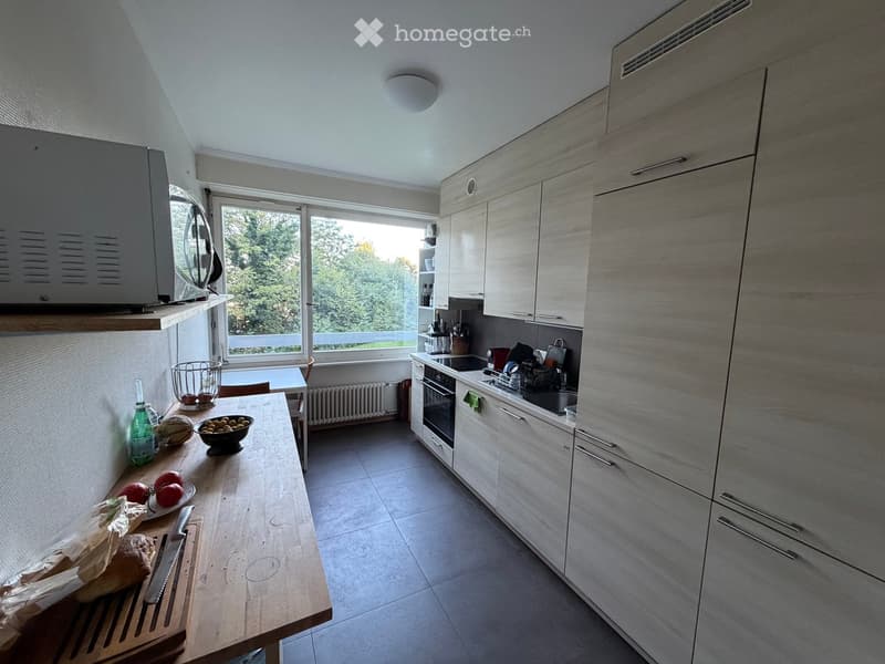 Modern kitchen with light wood cabinets, stainless steel appliances, and a large window overlooking greenery. The kitchen island has a wooden countertop with fruits, a bowl, and a bottle. The stove area includes an oven, microwave, and sink.