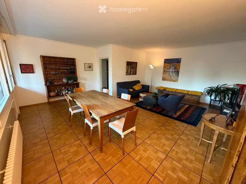 A well-lit living and dining area with wooden flooring, a sofa, a dining table with chairs, a cabinet, and a floor lamp.