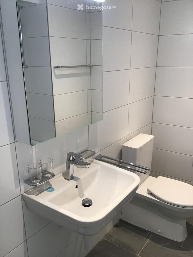 Modern bathroom with a white sink, chrome faucet, toilet, and a large mirror. Two clear cups are placed on the sink counter.