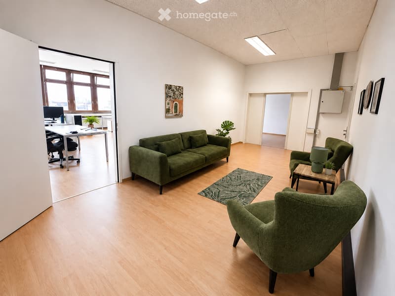 Modern living room with light wooden flooring, green sofa, armchairs, small table, lamp, and a plant. Large windows provide natural light, and a small office area is visible through an open doorway.