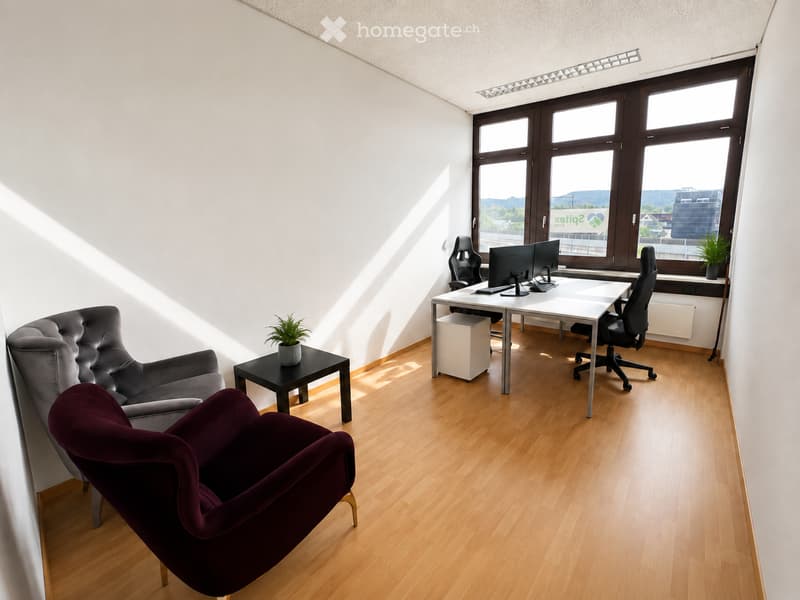 Modern office space with wooden flooring, large windows, a grey sofa, a dark red armchair, a small black side table with a plant, a white desk with two computer screens, and two office chairs.