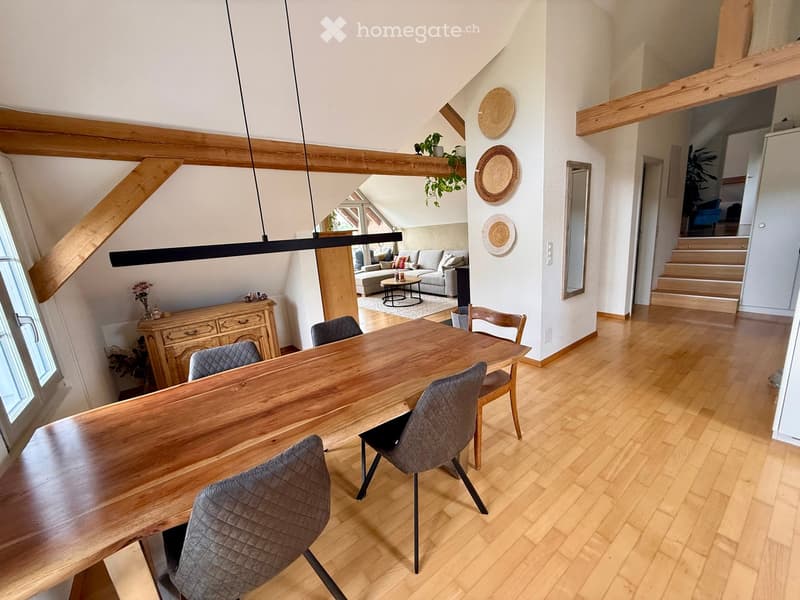 Open-plan dining and living area with wooden floors, a long wooden dining table with four chairs, a grey sofa, a coffee table, and a modern pendant light fixture.