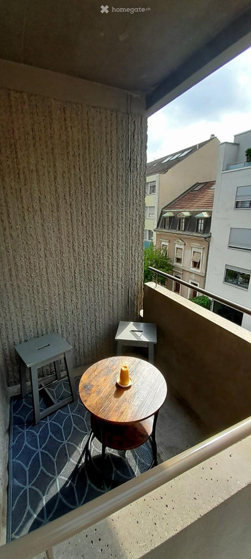 Balcony with a round wooden table, two stools, and a small decorative item on the table. The balcony has a textured wall and a patterned rug on the floor.