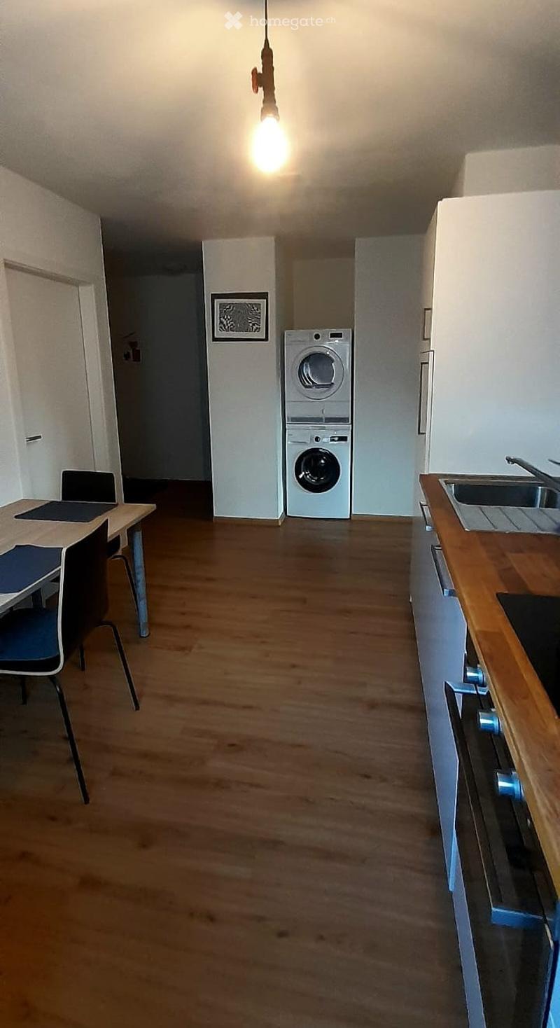 Open-plan room with wooden flooring, a small dining area with a table and chair, a laptop, and a stacked washing machine and dryer unit.