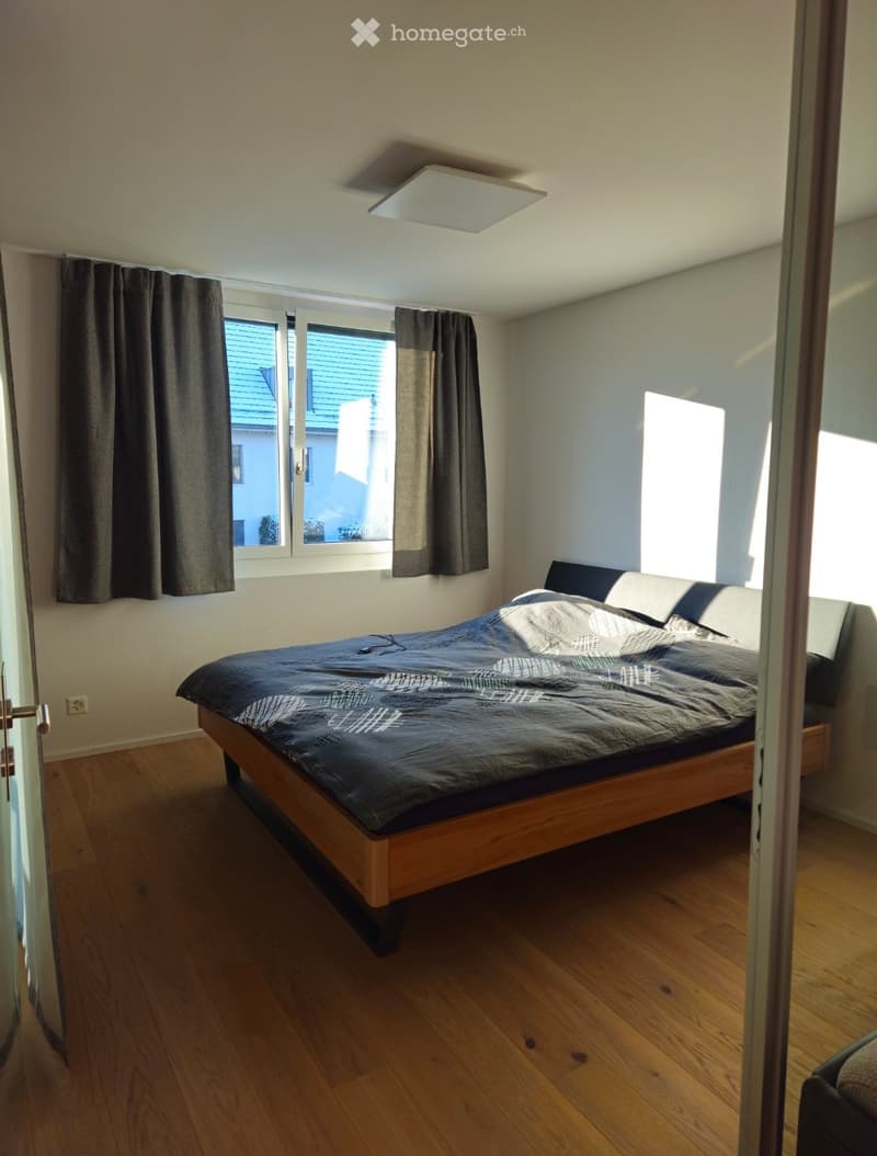 A small, well-lit bedroom with wooden flooring, a single bed with dark bedding, and dark curtains on the windows.