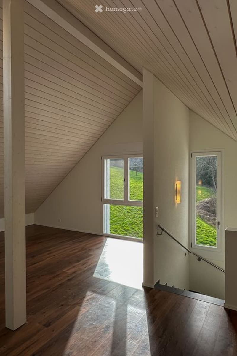 Bright, empty attic room with wooden floors, slanted wooden ceiling, large windows offering a view of a green lawn, and a staircase leading down.