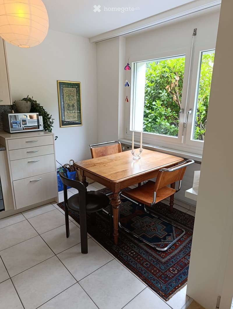 Bright room with a wooden dining table, chairs, microwave, cabinet, and a framed picture on the wall. Natural light from large windows and a view of greenery outside.