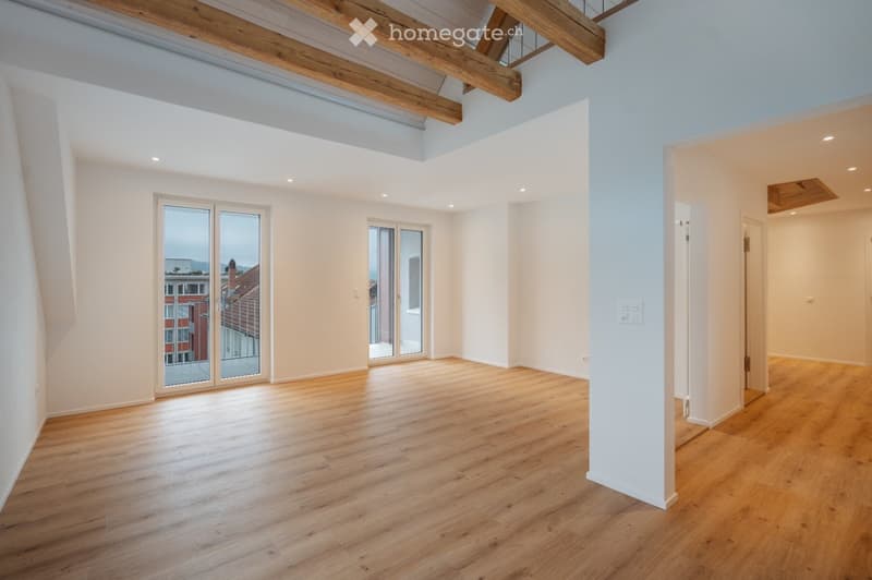 Empty room with wooden flooring, white walls, exposed wooden beams on the ceiling, and large glass doors leading to a balcony with a city view.