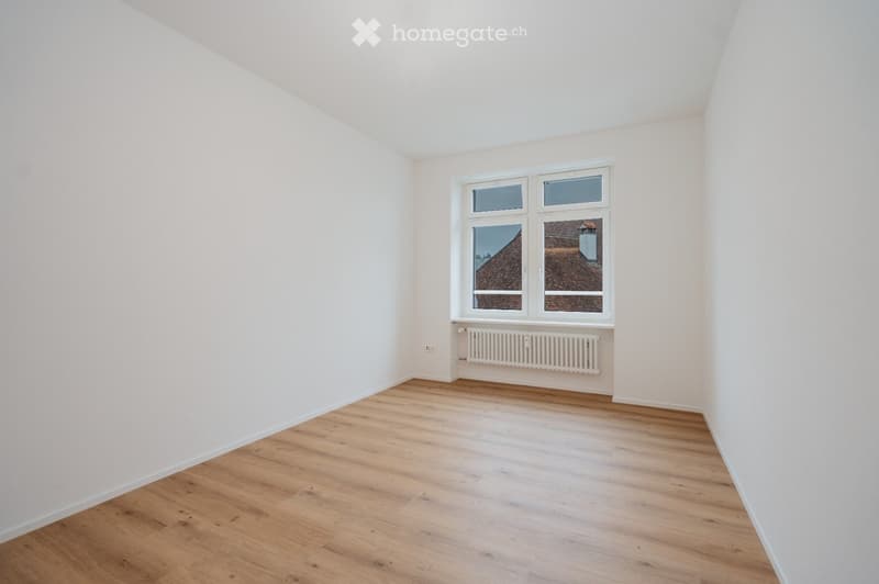 Empty room with light wooden flooring, white walls, and a large window with white frame showing a view of a roof and chimney.