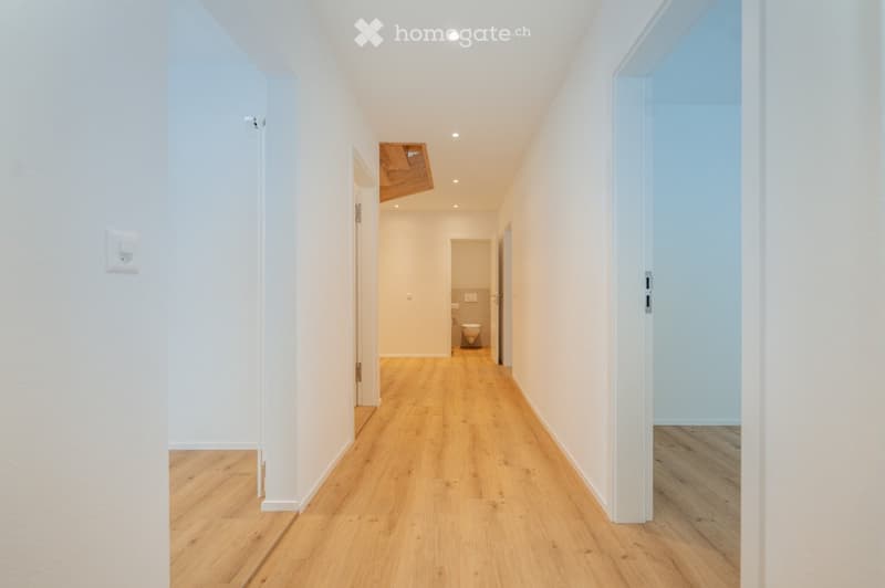 A clean, modern hallway with light wooden flooring and white walls, leading to a small bathroom with a toilet and sink.