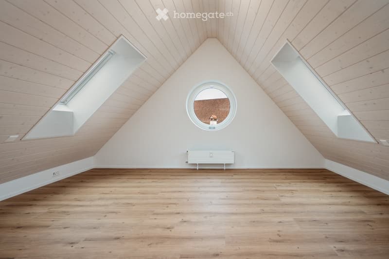 Empty attic room with wooden floors, slanted wooden ceiling, skylights, and a round window showing a view of the roof.