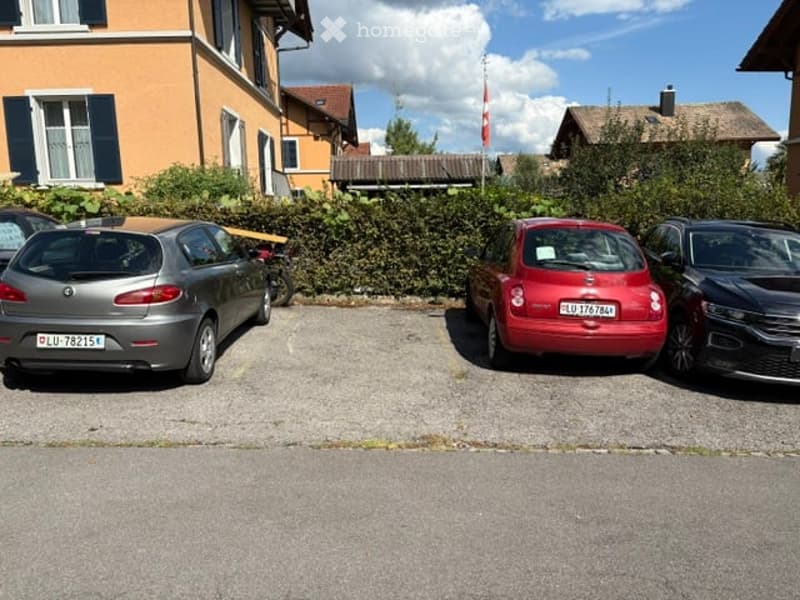 Residential building with a parking area in front, several cars parked, and a hedge separating the parking lot from the building.