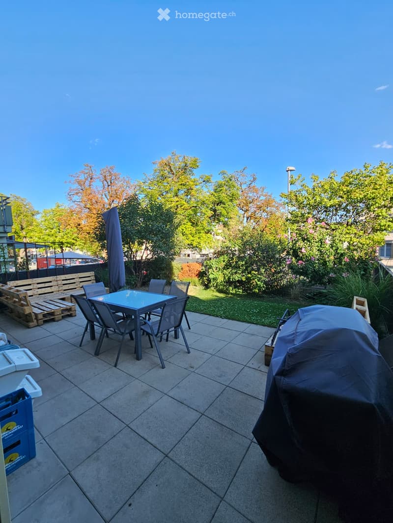 Outdoor patio with tiled floor, a table and chairs, a covered grill, a bench, and surrounding greenery.