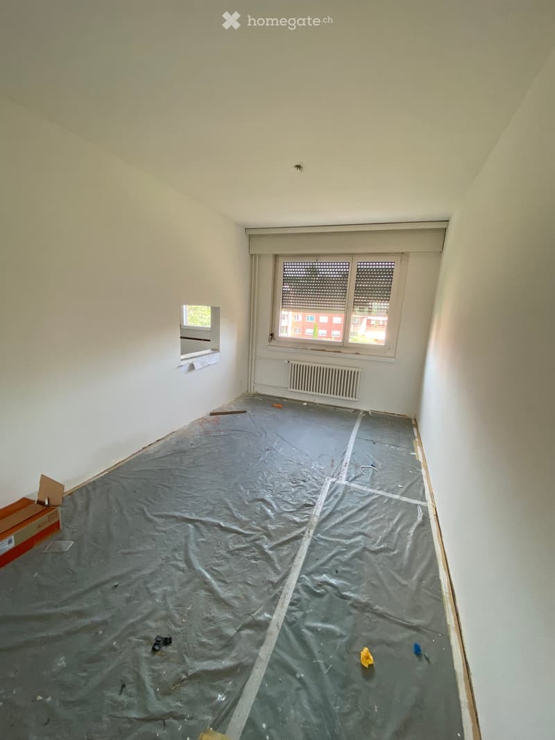 Empty room with bare walls, a window, and a radiator. The floor is covered with a protective plastic sheet, and there are some scattered items and debris.