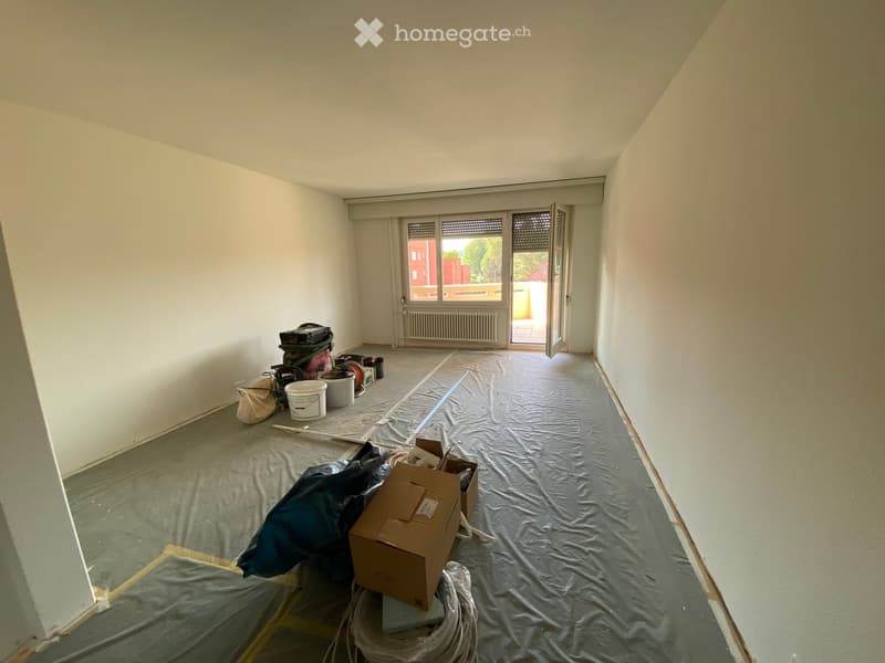 Empty room with bare walls and a covered floor, showing renovation materials and tools scattered around.