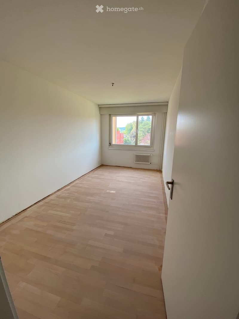 Empty room with wooden flooring, white walls, and a window with a view of trees and a building outside.