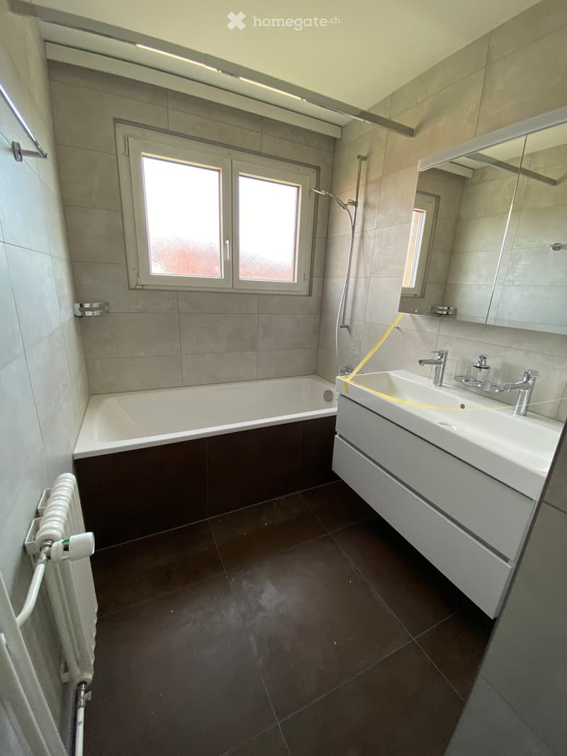Modern bathroom with a bathtub, sink, and mirror. Features dark tiled flooring and a window with frosted glass.