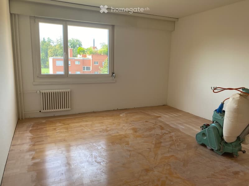Empty room with wooden flooring, large window, radiator, and a floor sanding machine in the corner.
