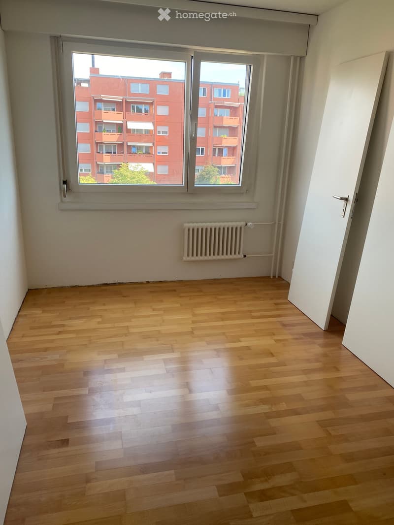 Empty room with wooden flooring, white walls, a radiator, and a large window overlooking a red apartment building.