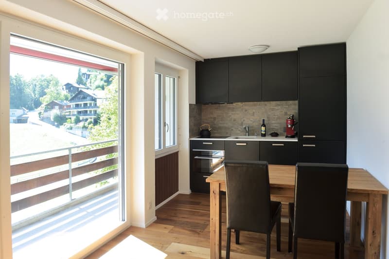Modern kitchen with dark cabinets, wooden floor, large window with view, dining table with chairs, sink, oven, and various items on counter.