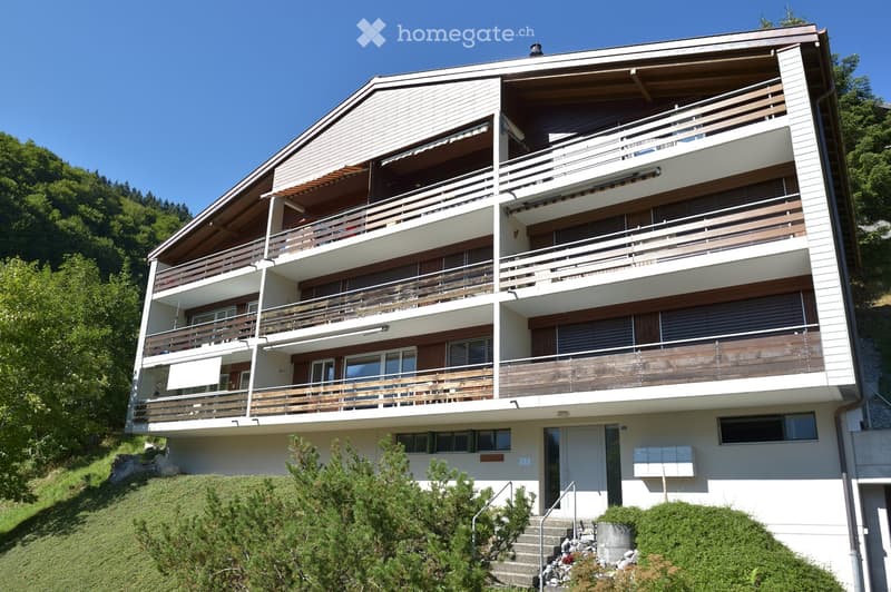 A modern, multi-story residential building with white walls, wooden balconies, and large windows, situated on a hillside with greenery in the background.