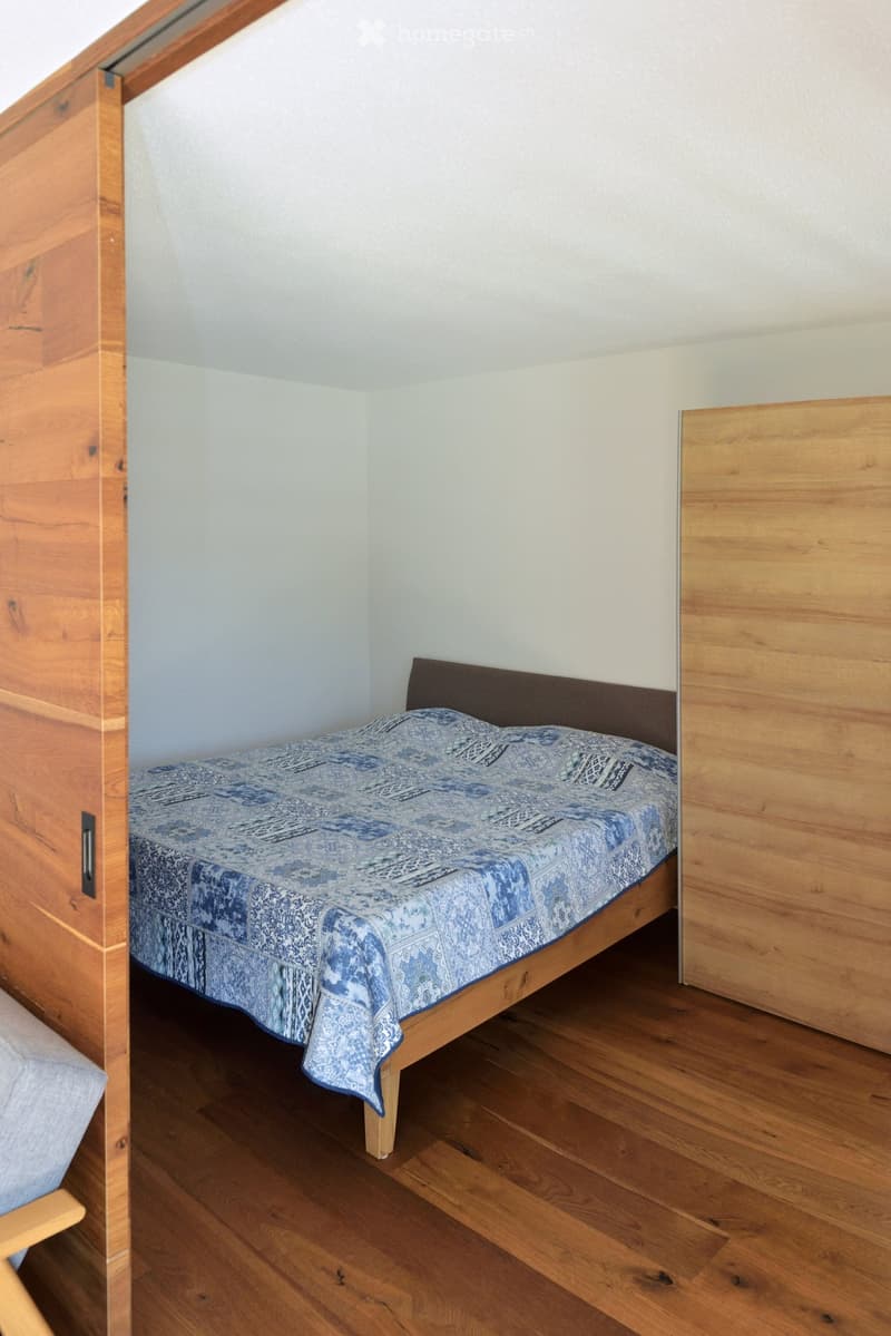 A small bedroom with a wooden bed, blue patterned bedspread, and wooden headboard. The room features light-colored walls and wooden flooring.