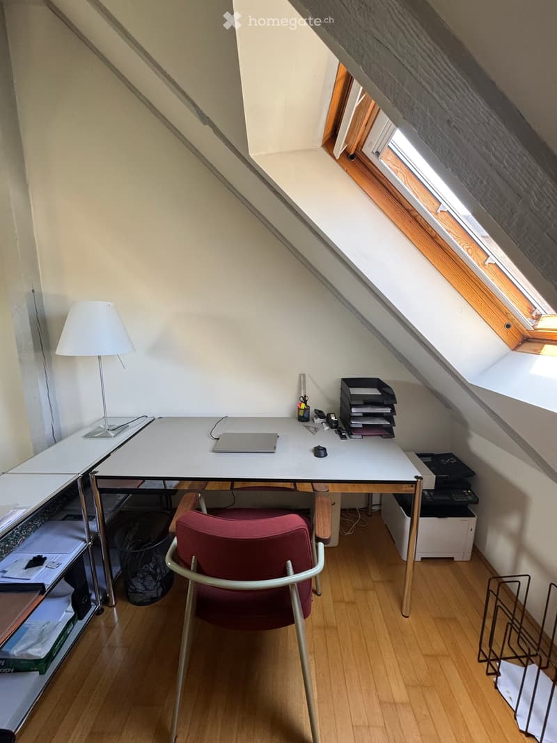 A small, well-lit attic office with wooden flooring, a white desk, a red chair, a lamp, a laptop, and various office supplies.
