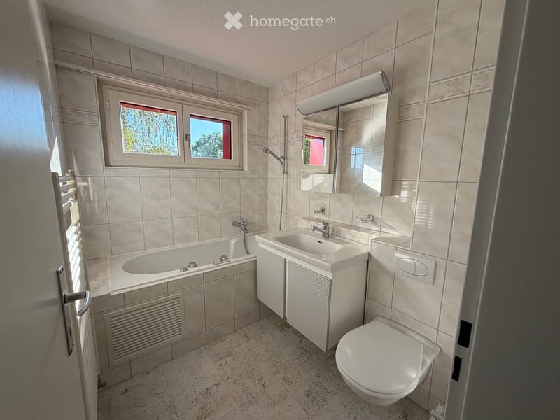 Modern bathroom with a bathtub, sink, toilet, and mirror. Features tiled walls and a window with a view of greenery.