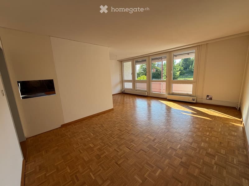 Empty room with wooden herringbone flooring, large windows, and white walls.