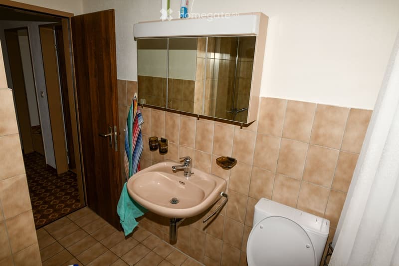 Bathroom with a sink, mirror, toilet, and a towel hanging on the wall. The sink has a chrome faucet and a small shelf above it. The walls are tiled in beige, and the floor is tiled in a patterned design.