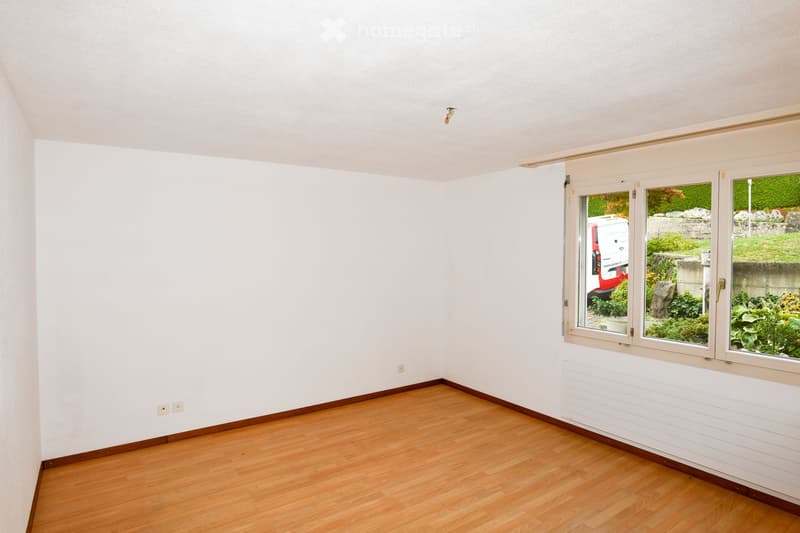 Empty room with wooden flooring, white walls, and large windows showing a view of greenery and a parked vehicle.