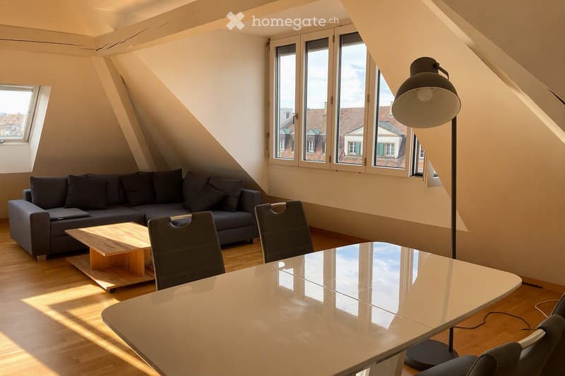 Bright attic living space with wooden flooring, large windows, a grey sofa, a wooden coffee table, a glass dining table, and chairs.