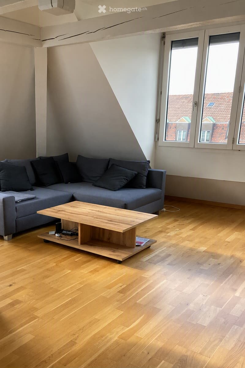 Modern living room with a grey sectional sofa, wooden coffee table, and hardwood flooring. Large windows offer a view of the city.
