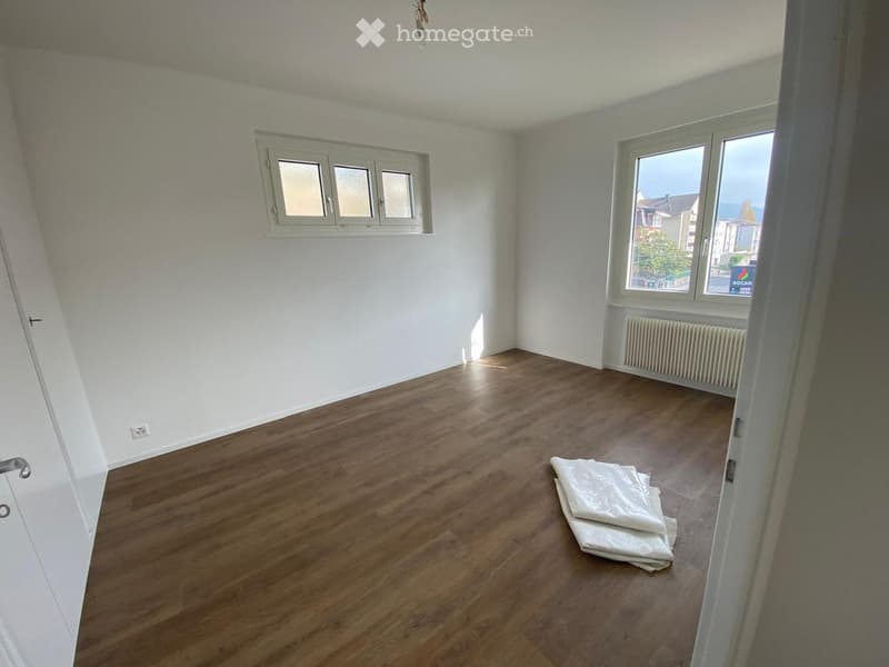 Empty room with wooden flooring, white walls, and two windows. A radiator is visible under one window, and a folded white item is on the floor.