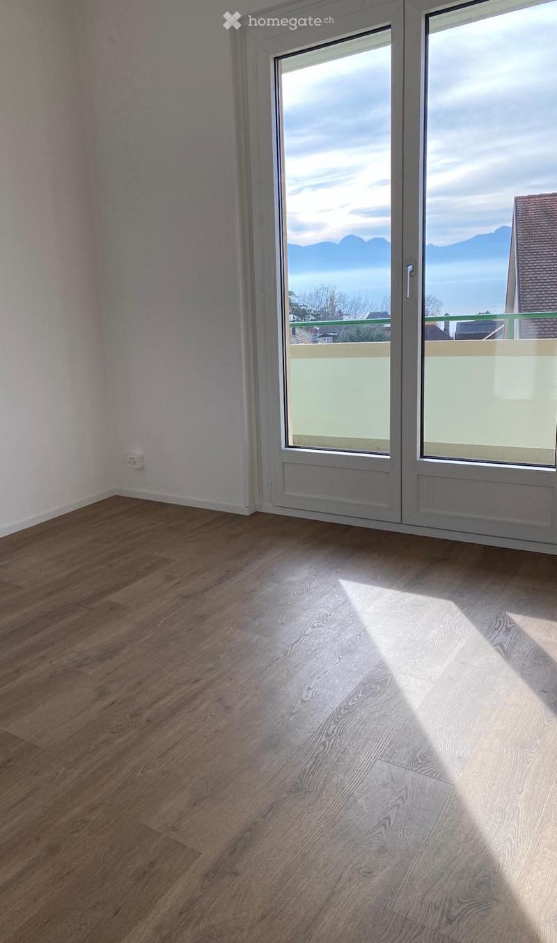 Empty room with wooden flooring and large windows offering a scenic view of mountains and a lake.