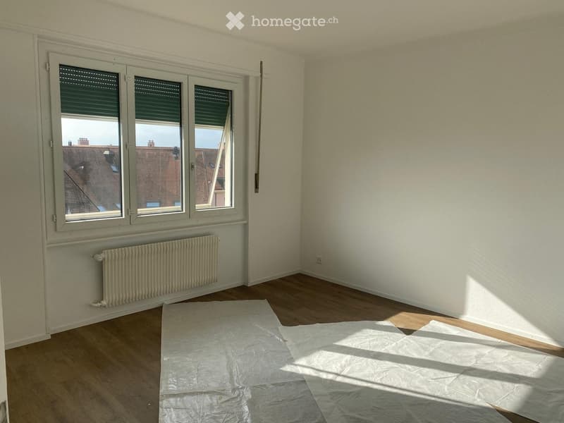 Empty room with wooden flooring, white walls, large windows with blinds, radiator, and protective plastic sheets on the floor.