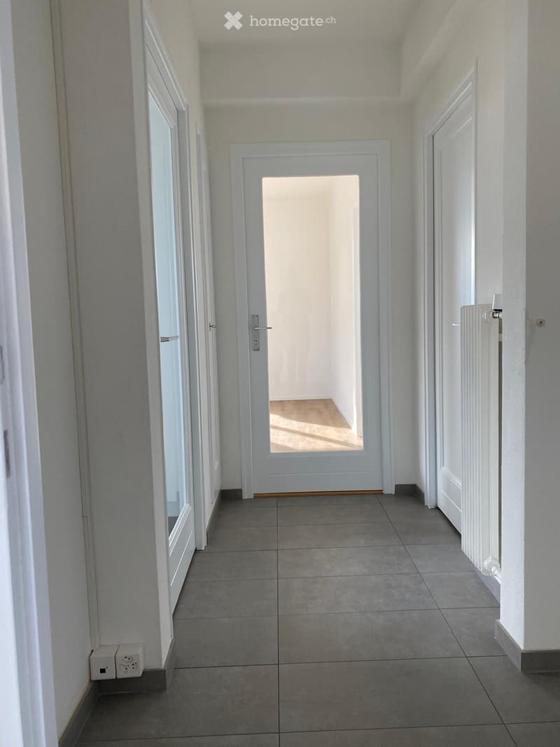 Empty hallway with tiled floor, white walls, and multiple doors including a glass-paned door at the end.