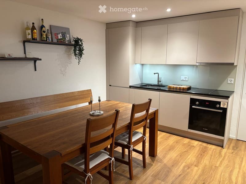 Modern kitchen with light-colored cabinets, a wooden dining table with four chairs, a built-in oven, and a sink with a faucet. A shelf holds bottles and a framed picture, and a small plant is visible.