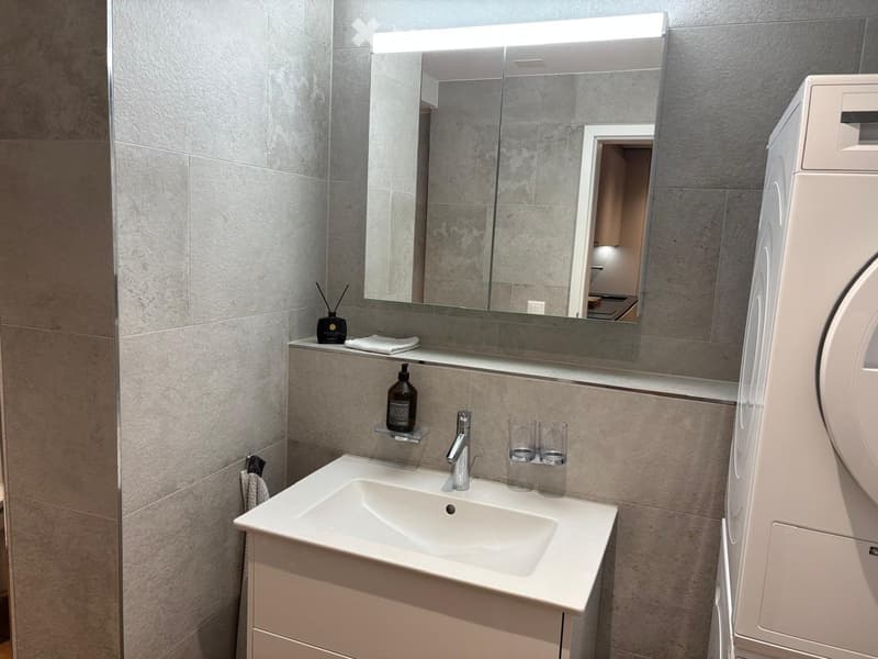 Modern bathroom with a white sink, chrome faucet, large mirror with built-in lighting, and a towel rack. A small bottle and two cups are placed on the sink counter.
