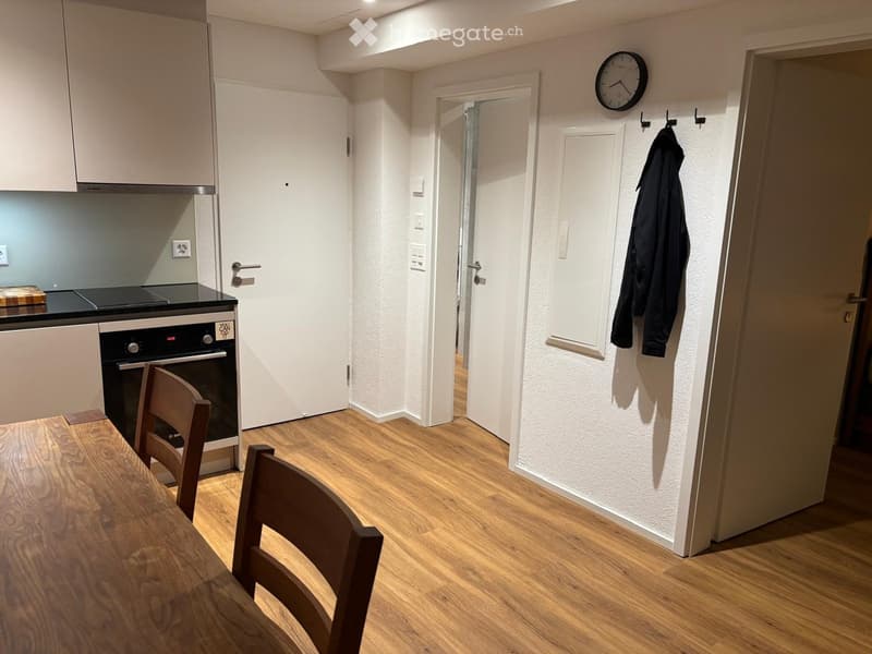 A modern, compact kitchen area with light wooden flooring, white walls, and built-in cabinets. A small dining table with chairs is visible, along with an oven and a clock on the wall.