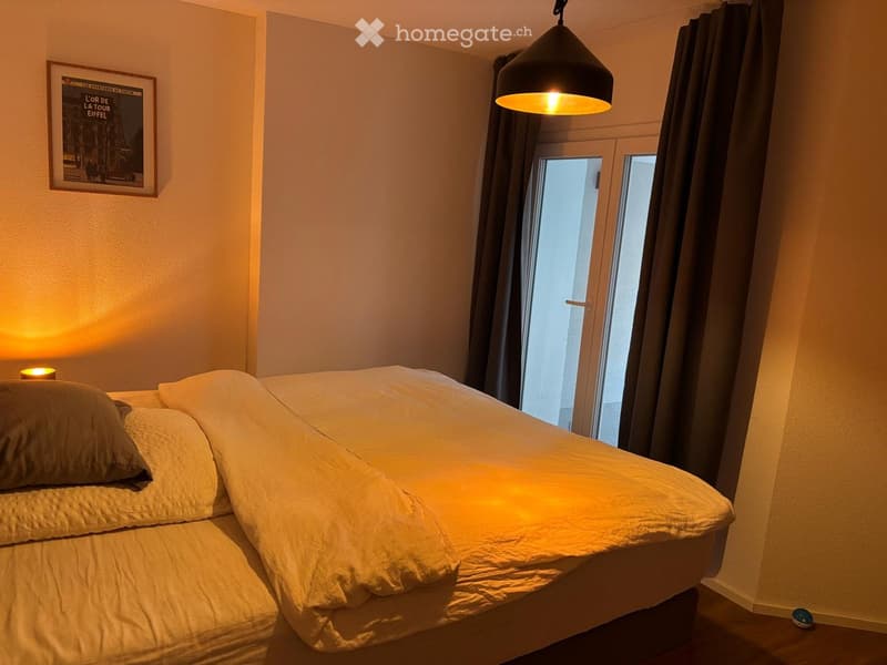 Cozy bedroom with a neatly made double bed, warm lighting from a hanging lamp and bedside lamp, large window with dark curtains, and a framed picture on the wall.