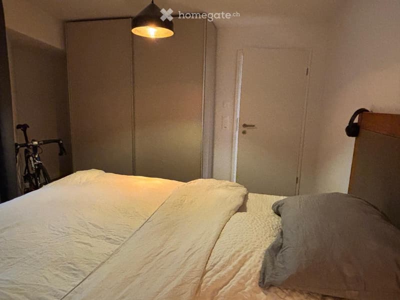 Bedroom with a neatly made bed, a bicycle leaning against the wall, and a hanging light fixture above the bed.